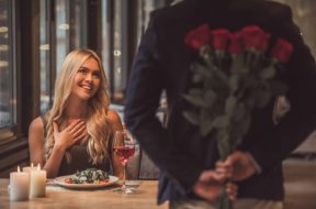 Beautiful girl is sitting at the table in restaurant and smiling while a man is holding roses behind his back