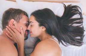 Cheerful and nice couple is lying in bed close to each other. Girl holds her hand on his neck. They are touching their noses. This picture is very nice and tender
