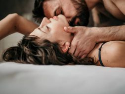 Side view photo of a beautiful couple in bed, being gentle and sensual. Handsome guy with a beard on top of the beautiful brunette is kissing her neck while her eyes are closed. Making love concept