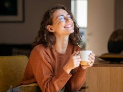 Portrait of joyful young woman enjoying a cup of coffee at home. Smiling pretty girl drinking hot tea in front of the window in winter. Excited woman wearing spectacles and sweater and laughing in an autumn day.