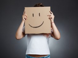 Young beautiful girl hiding head behind carton with smiley over grey background. Copy space.