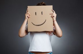Young beautiful girl hiding head behind carton with smiley over grey background. Copy space.
