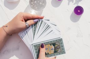 Hands with purple nails and rings hold deck of Tarot cards on white surface with crystal ball, candles and stones. Top view. Minsk, Belarus - 28.07.2021