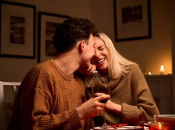 Happy young couple in love hugging, laughing, drinking wine, enjoying talking, having fun together celebrating Valentines day dining at home, having romantic dinner date with candles sitting at table.