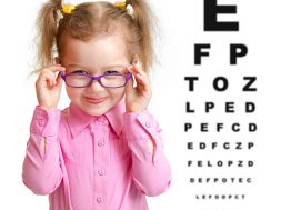 Smiling girl putting on glasses with blurry eye chart behind her isolated on white