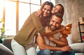 Happy dad and mom with their cute daughter and teddy bear hug and have fun sitting on the sofa in the living room at home.