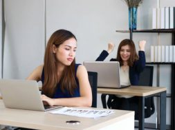 Young angry envious Asian business woman looking successful competitor colleague in office.