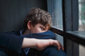 Pensive sad boy teenager with blue eyes in a blue shirt and jeans sitting at the window and closes his face with his hands.